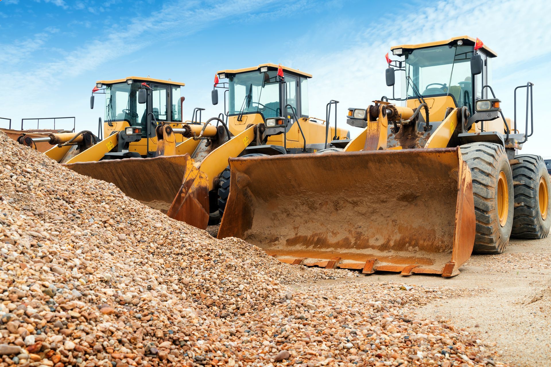 Three yellow construction loaders parked beside a pile of gravel under a blue sky.