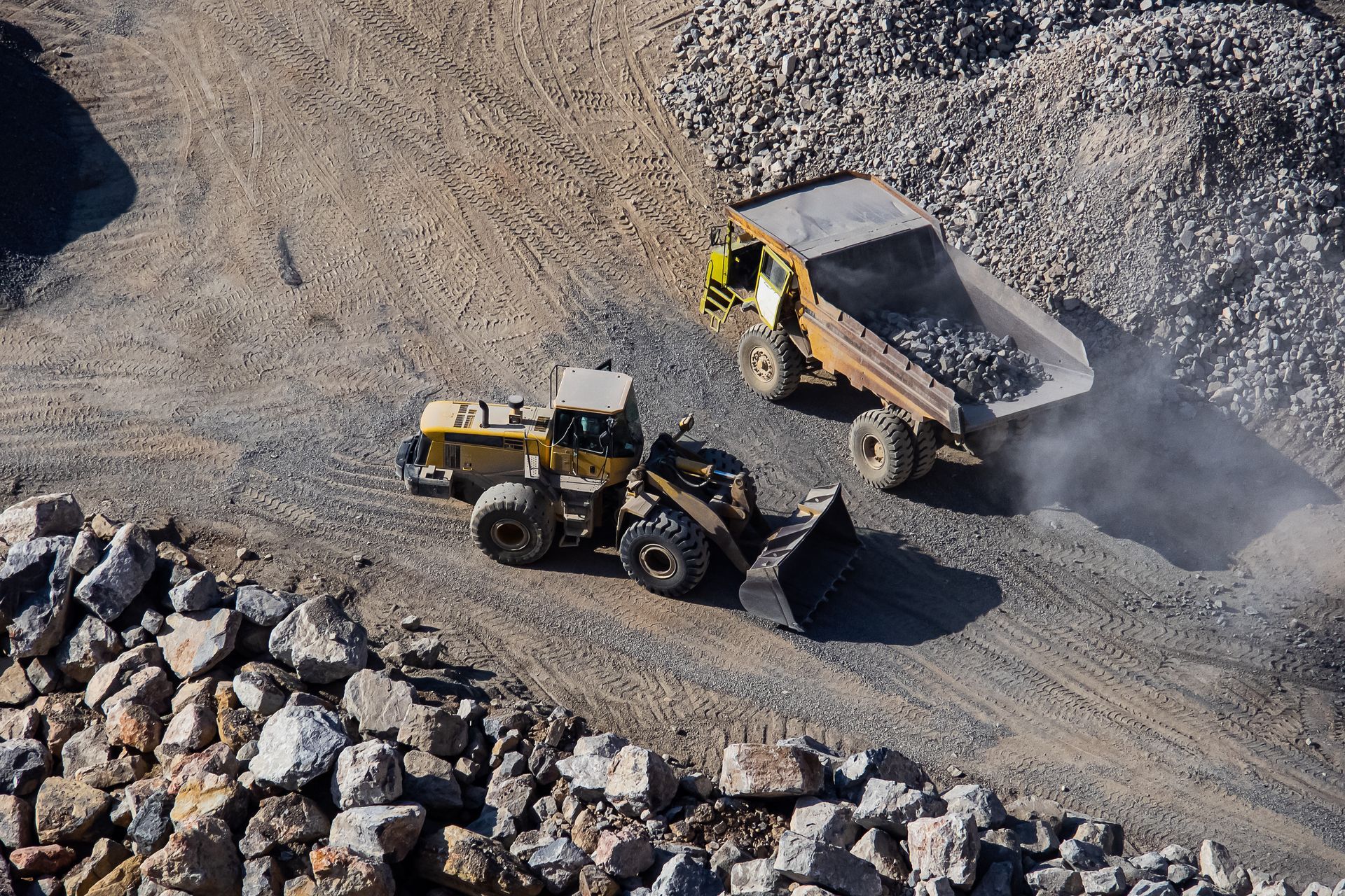 A yellow loader scoops rocks, loading a yellow dump truck in a quarry.