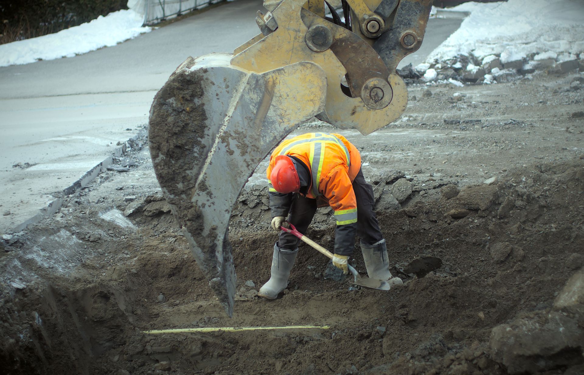 Construction worker digging in a hole, an excavator's bucket above him. He wears an orange vest, hard hat, and boots. Construction worker digging in a hole, an excavator's bucket above him. He wears an orange vest, hard hat, and boots.