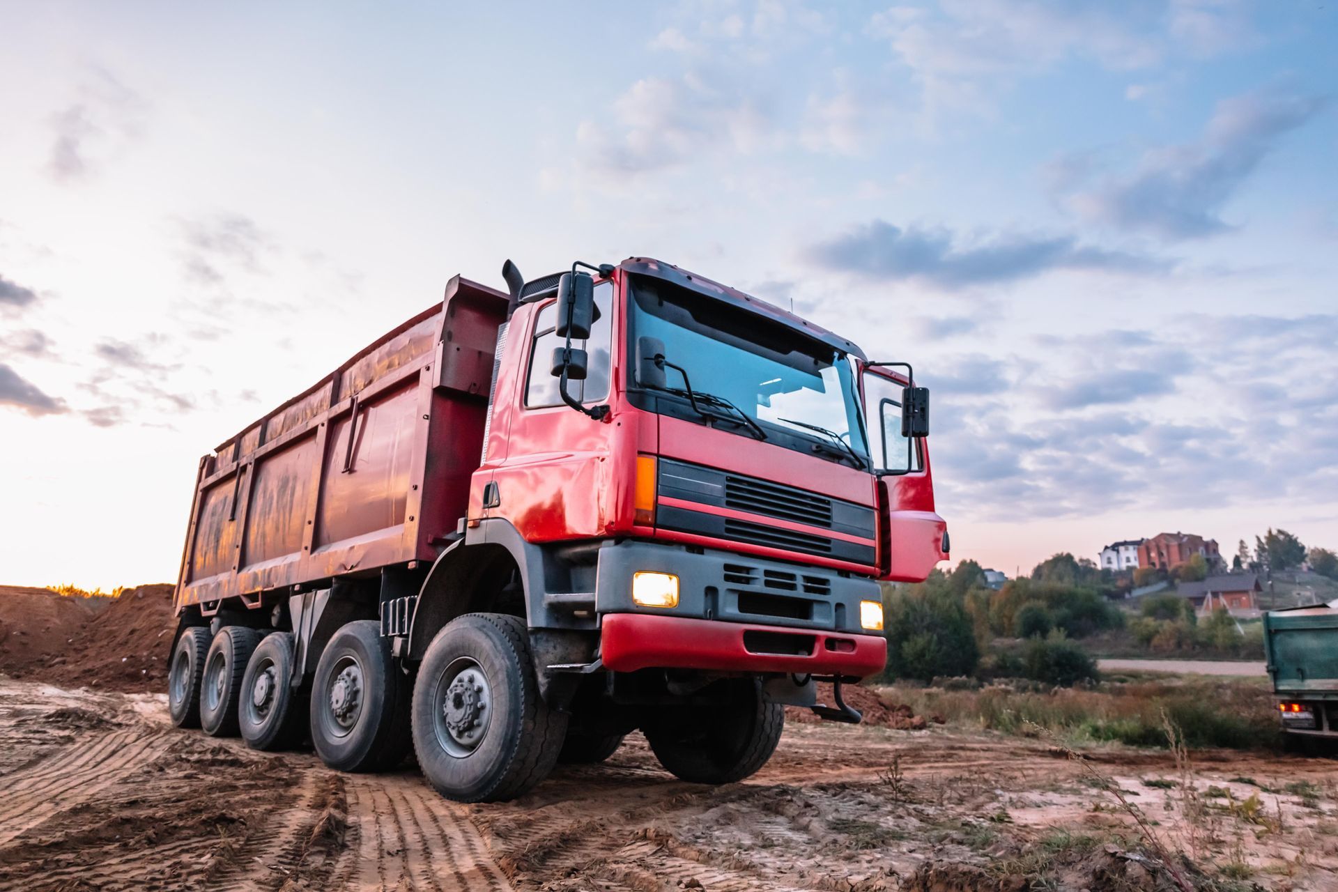 Red dump truck on a dirt road, likely construction site, under a cloudy sky.
