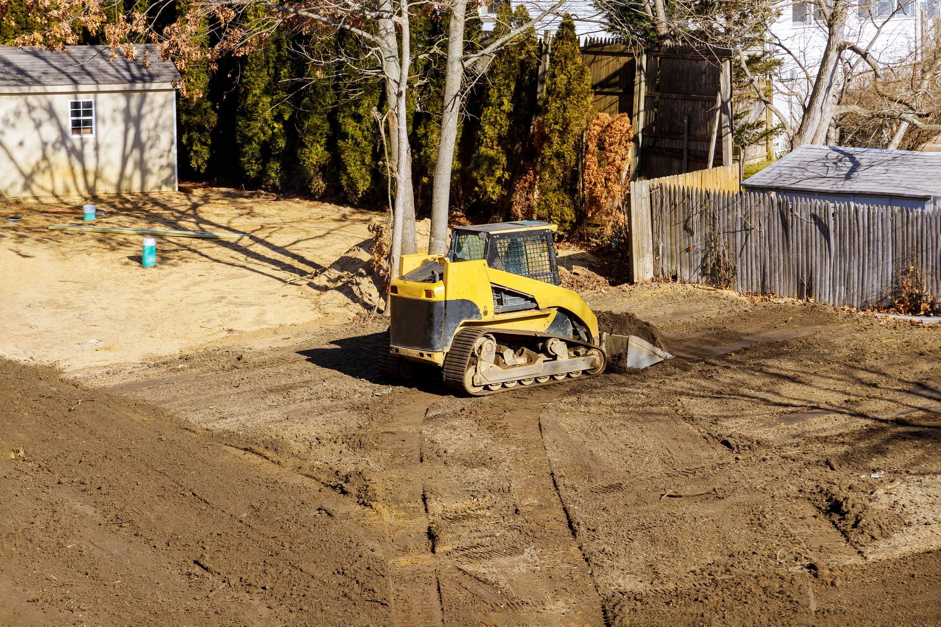 Yellow skid steer on a dirt lot, preparing the ground. Wooden sheds and fence in the background. Yellow skid steer on a dirt lot, preparing the ground. Wooden sheds and fence in the background.