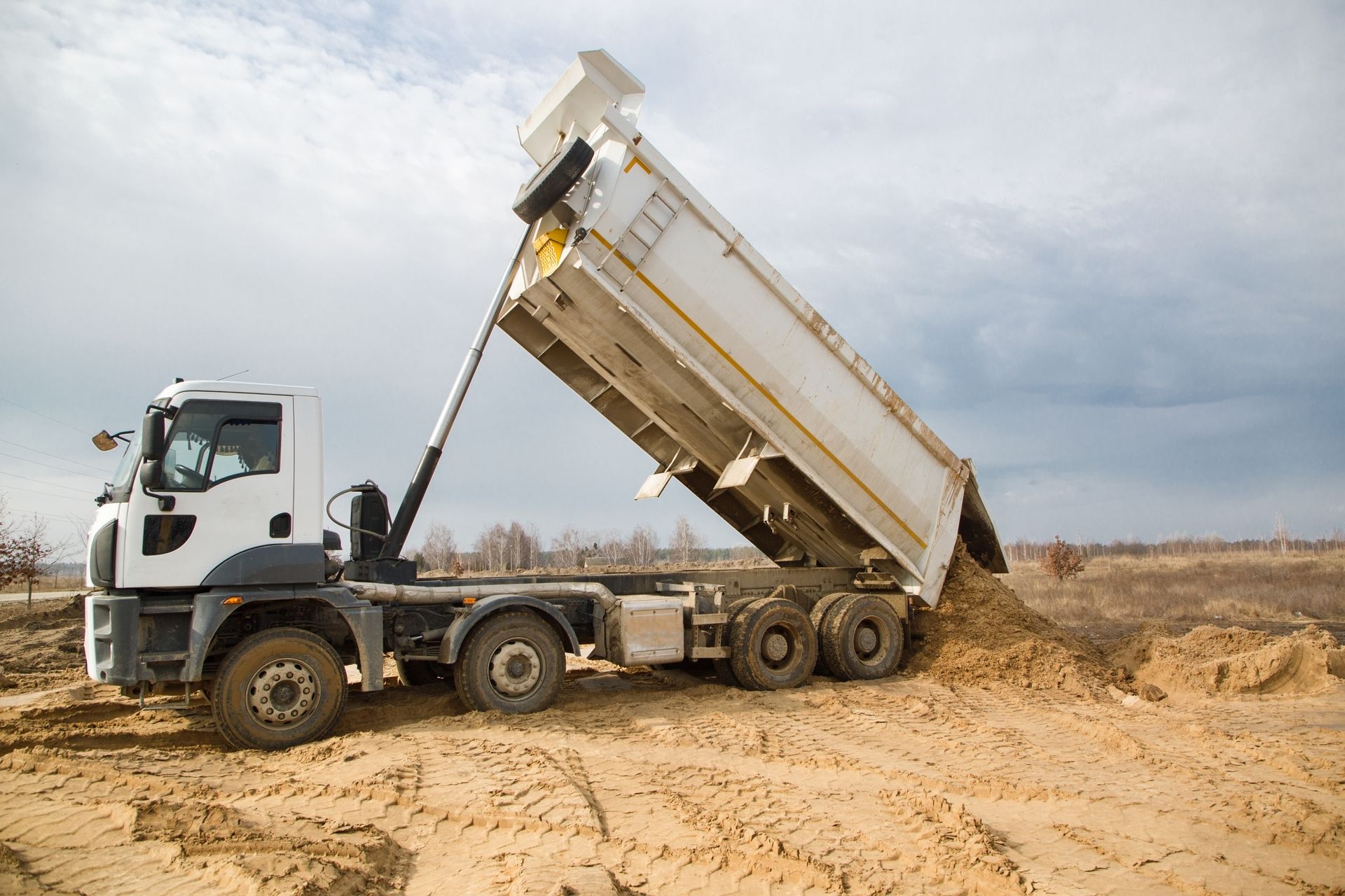 White dump truck unloading dirt on a sandy ground under a cloudy sky.