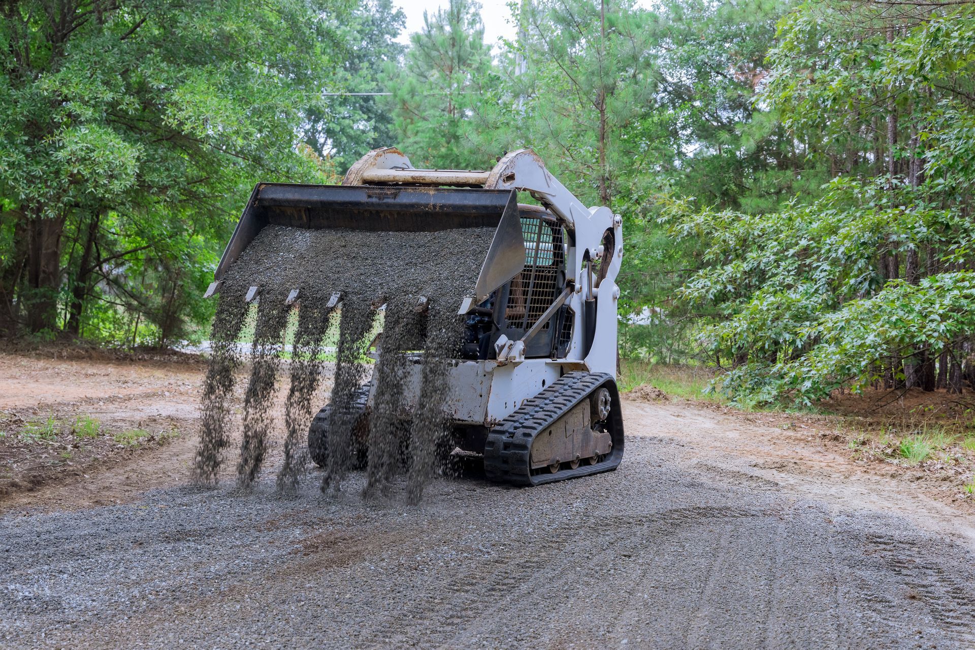 Skid steer dumping gravel onto a dirt road, trees in the background. Skid steer dumping gravel onto a dirt road, trees in the background.