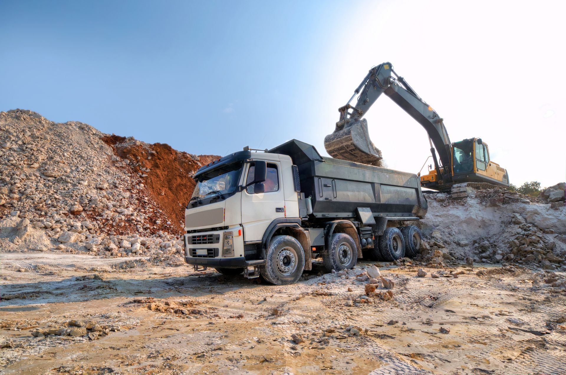 Excavator loading a dump truck at a construction site; dirt mounds in the background, blue sky.