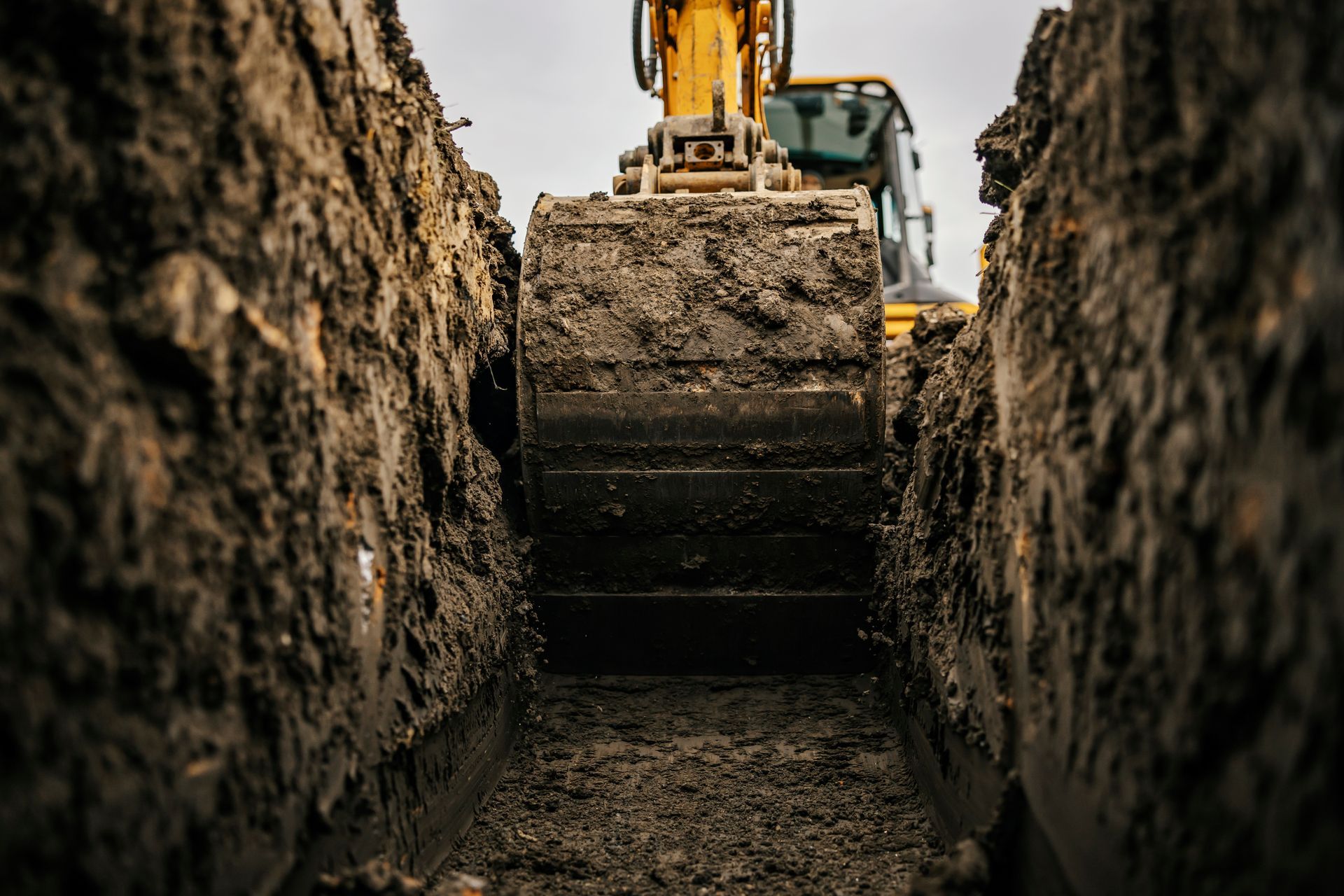 Excavator bucket in a narrow trench, scooping up dirt; construction site.