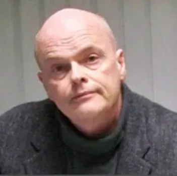 A bald man wearing a turtleneck and a jacket is looking at the camera.
