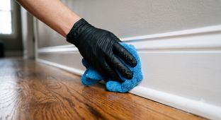 Man mops a wooden floor with a yellow bucket nearby while another person cleans a couch in the background.