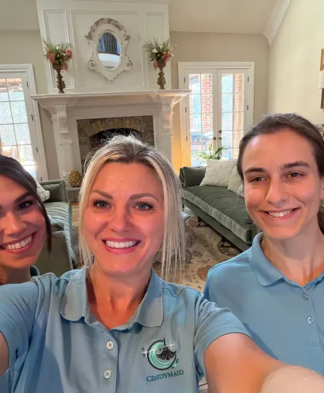 Three smiling people in blue polos taking a selfie in a bright living room with a fireplace.