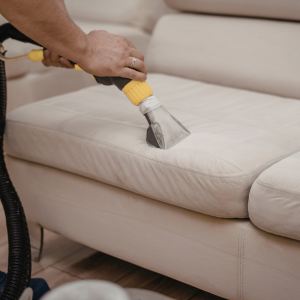 Person steam-cleans a beige couch with a handheld upholstery cleaner.