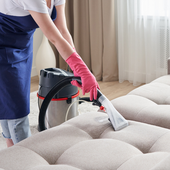 Cleaner vacuuming a beige sofa with a red-gloved hand in a bright living room