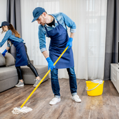 Man mops a wooden floor with a yellow bucket nearby while another person cleans a couch in the background.