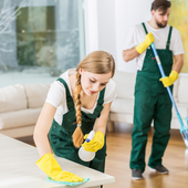 Two cleaners in green overalls and yellow gloves cleaning a bright living room with mop and cloth.