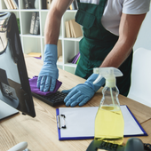 Person in green apron and blue gloves cleaning a keyboard with a spray bottle and cloth at a desk