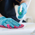 Gloved hands cleaning a white surface with a pink cloth and green spray bottle