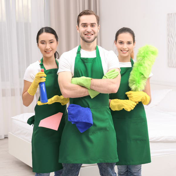 Three cleaners in green aprons holding cleaning supplies and cloths, posing in a bright room.