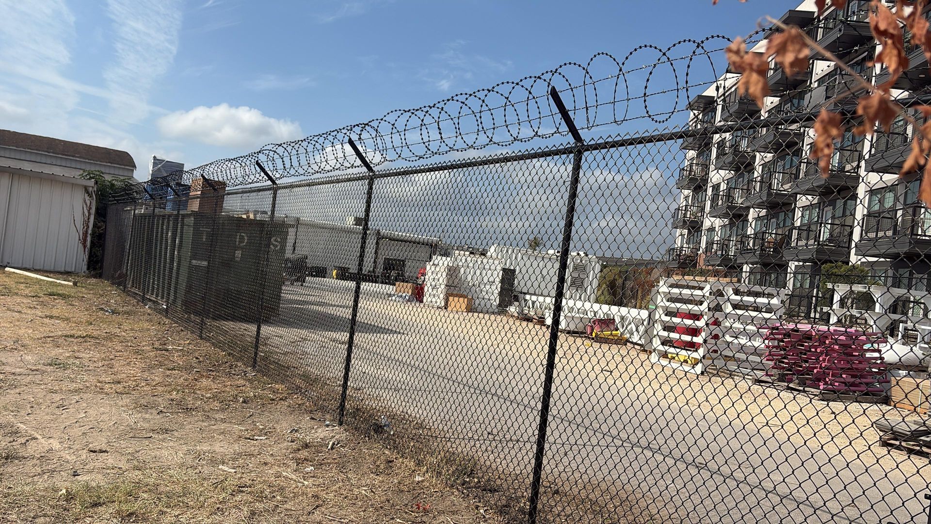 Chain-link fence topped with razor wire surrounds an industrial area with stacked materials and a white building under a blue sky.