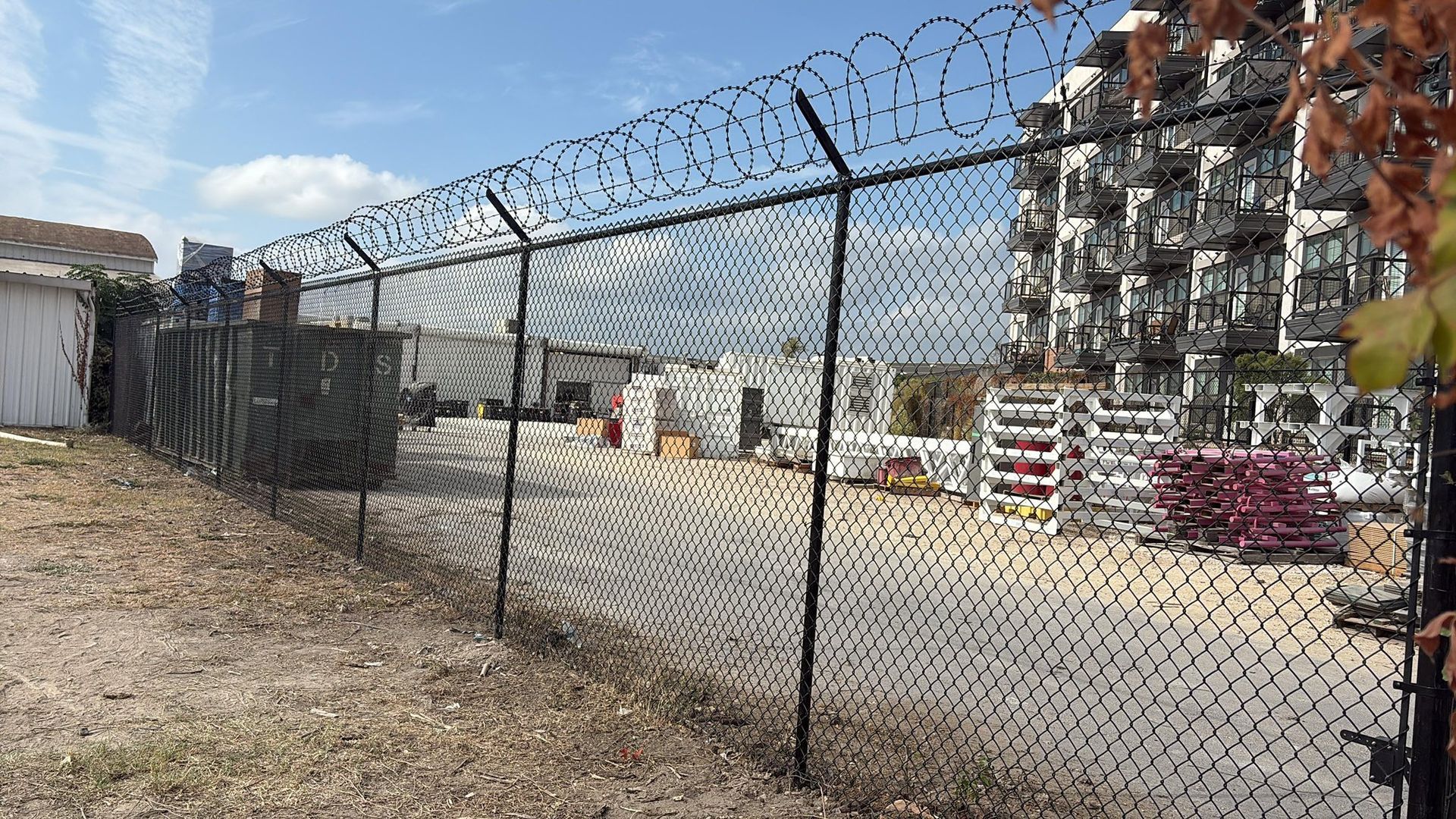 Black chain-link fence topped with barbed wire encloses an industrial area; buildings in the background.