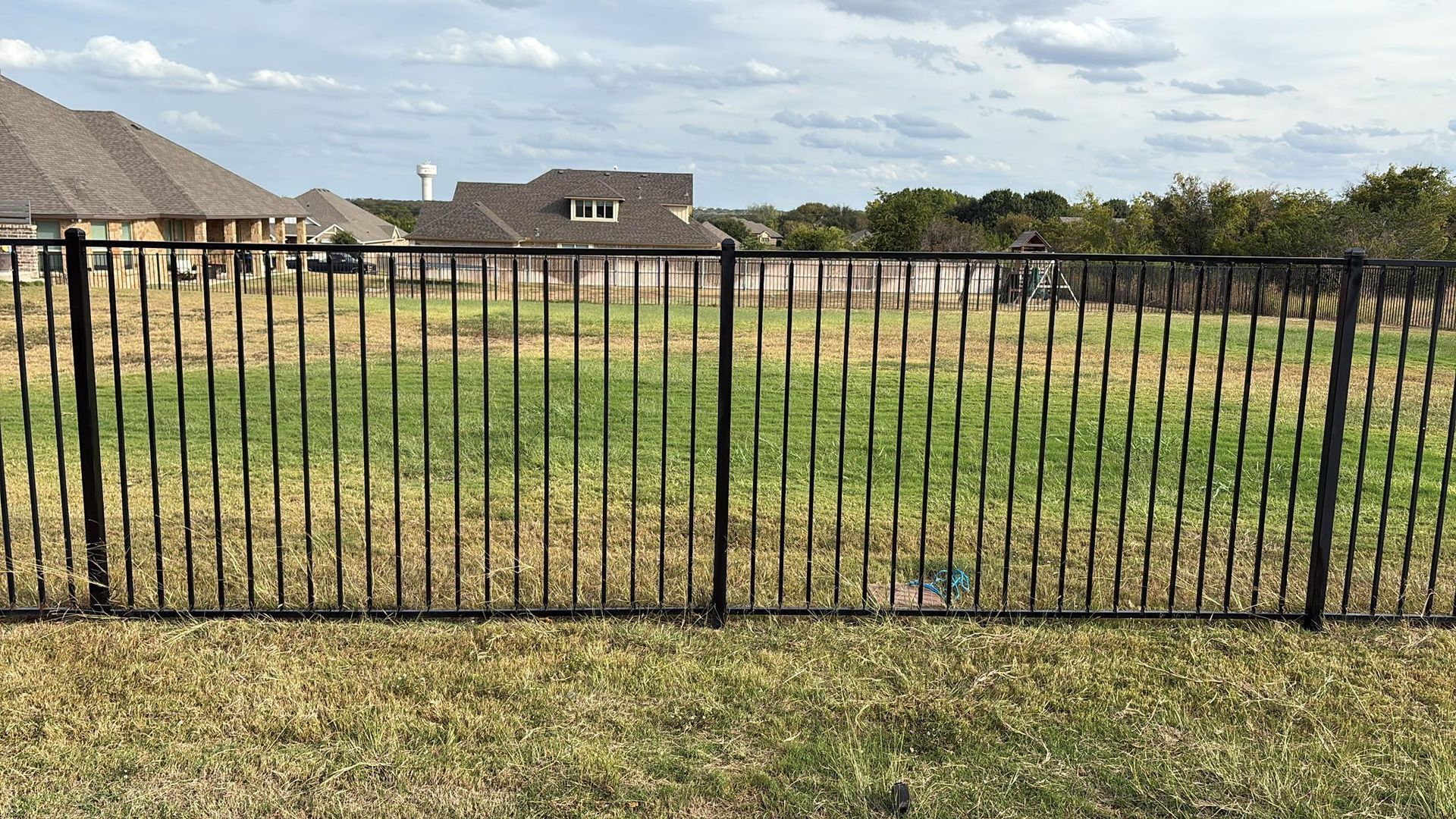 Black metal fence in front of a grassy field, houses in the background under a cloudy sky.