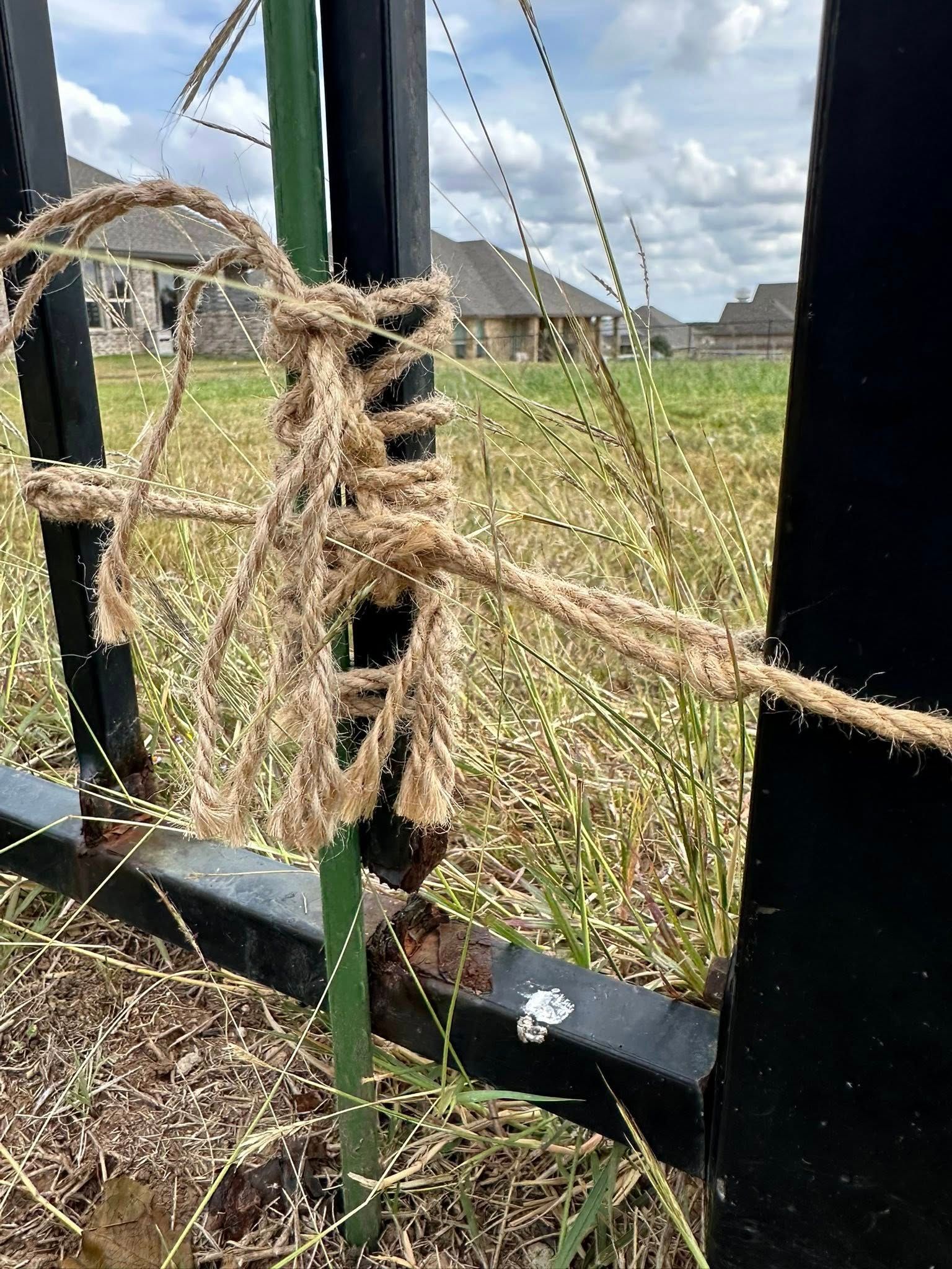 Jute rope tied around a black metal fence and green stake. Field and houses in the background.