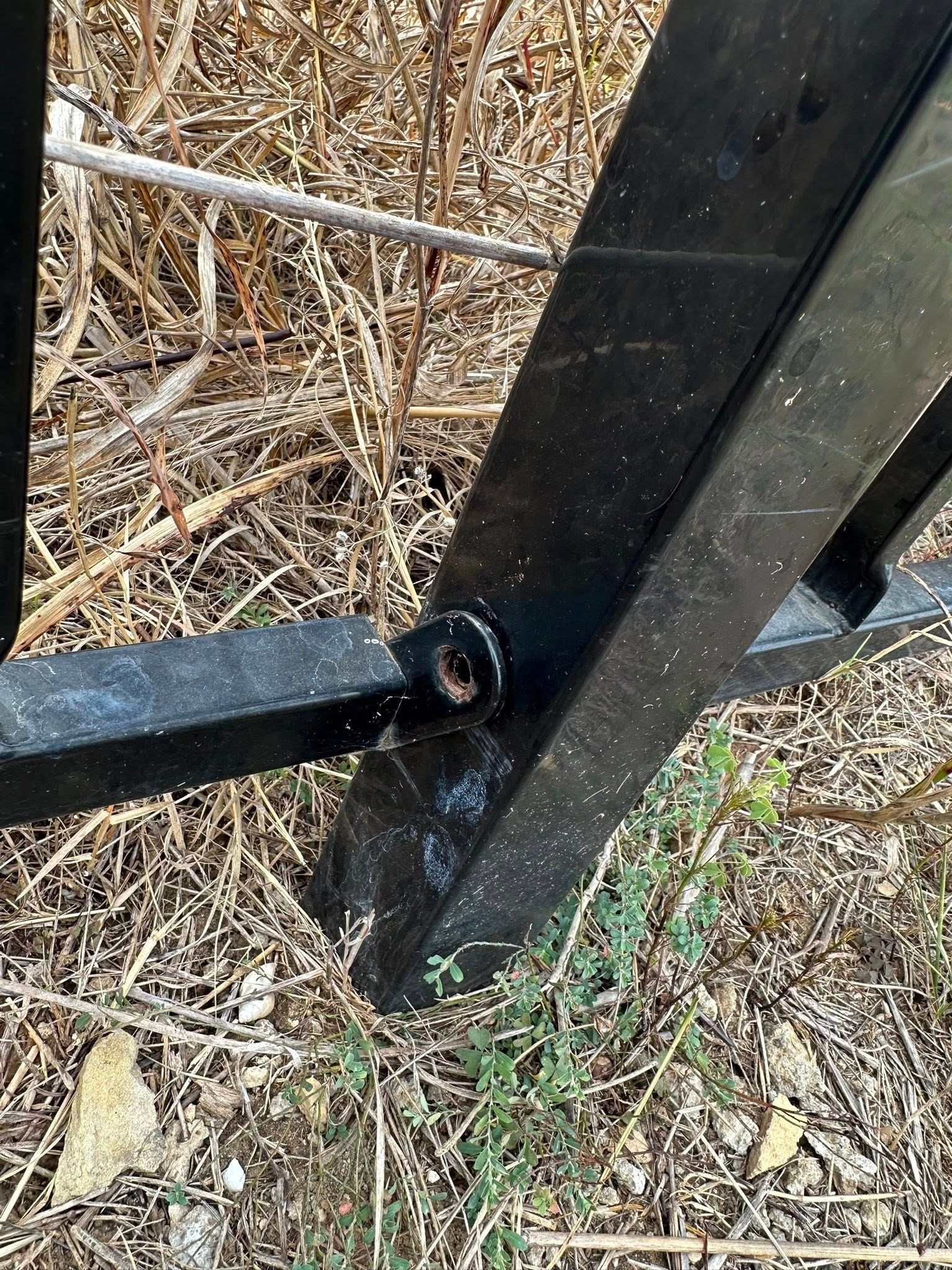 Black metal gate joint, worn paint, rusty bolt, dry grass backdrop.