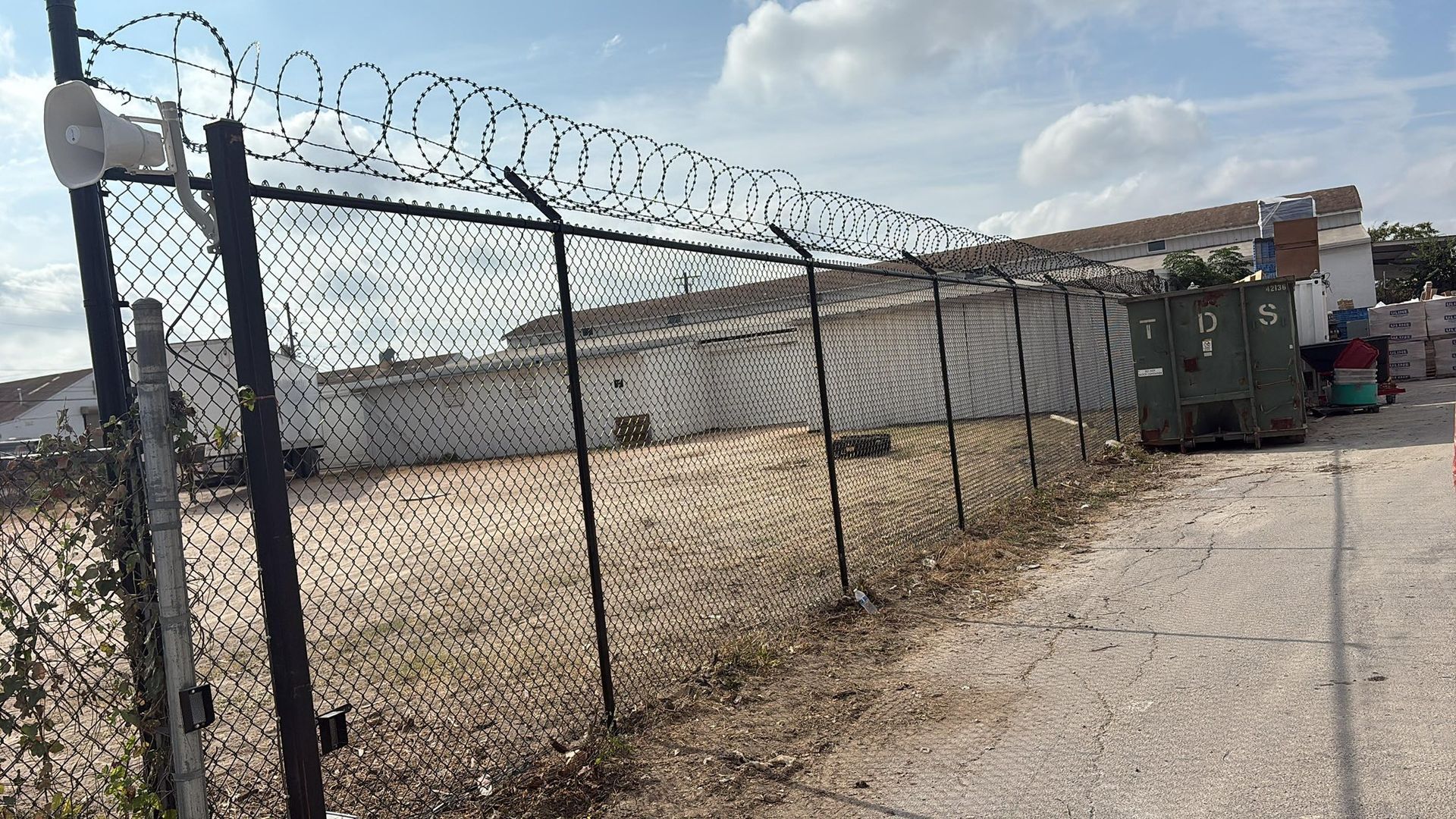 Black fence topped with barbed wire, gray building in background, gravel ground.