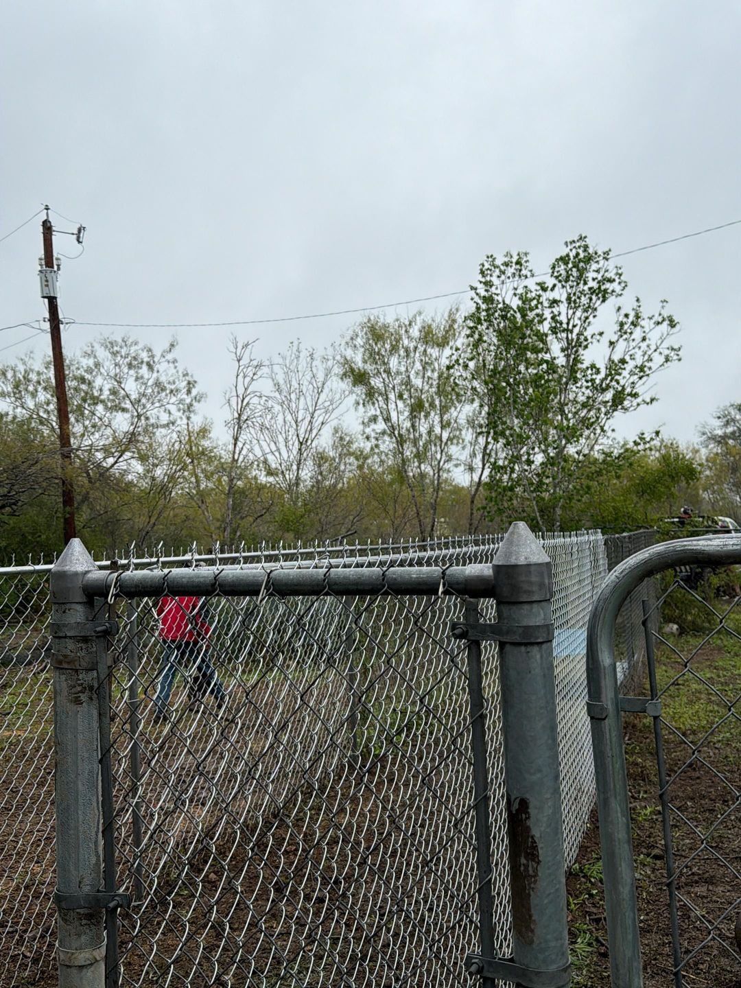 Chain-link fence with gate open, person in red jacket visible in the background, overcast day.