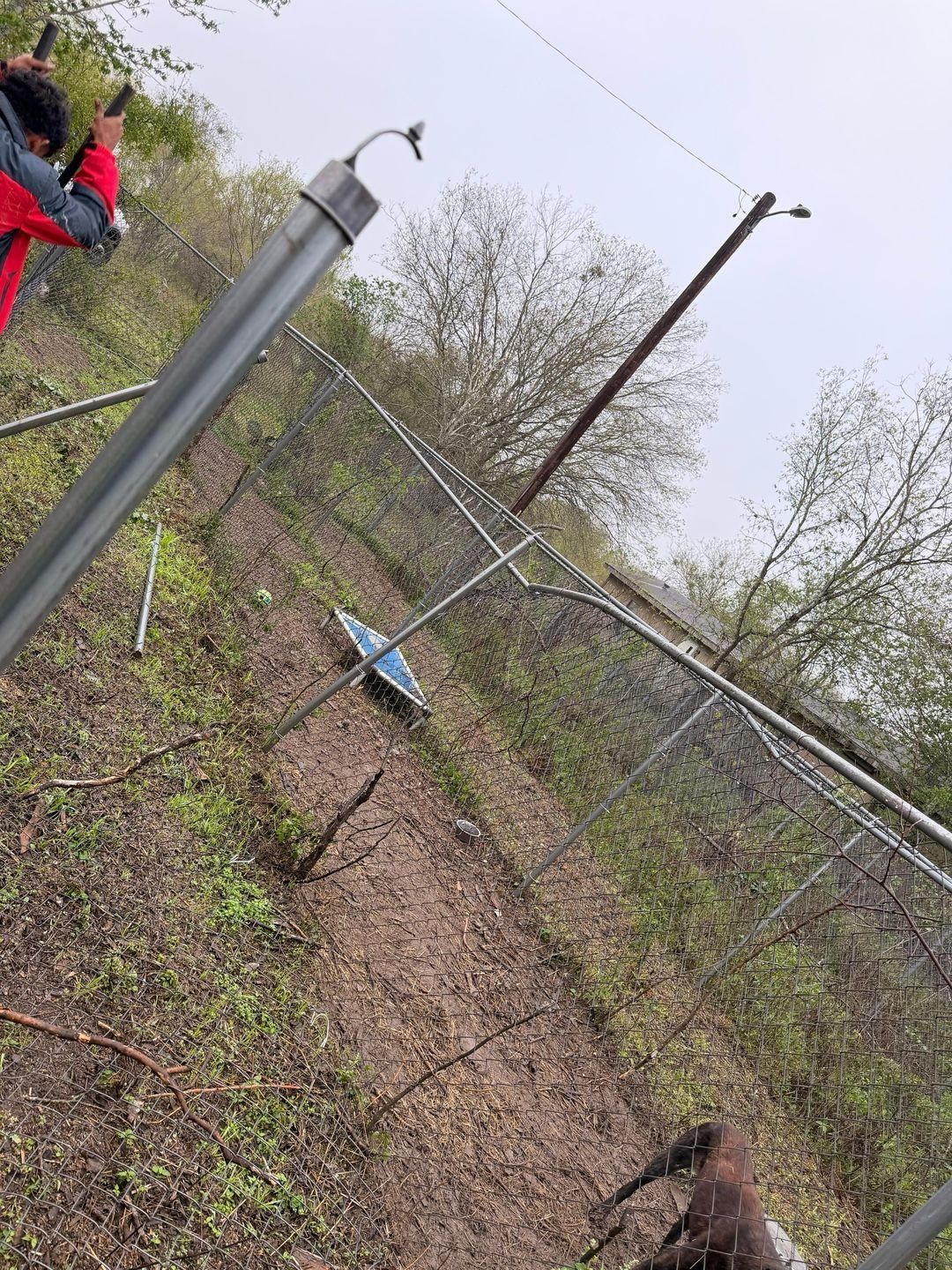 Fallen chain-link fence on a grassy hillside. Bent metal poles and wire are visible. Person in red jacket.