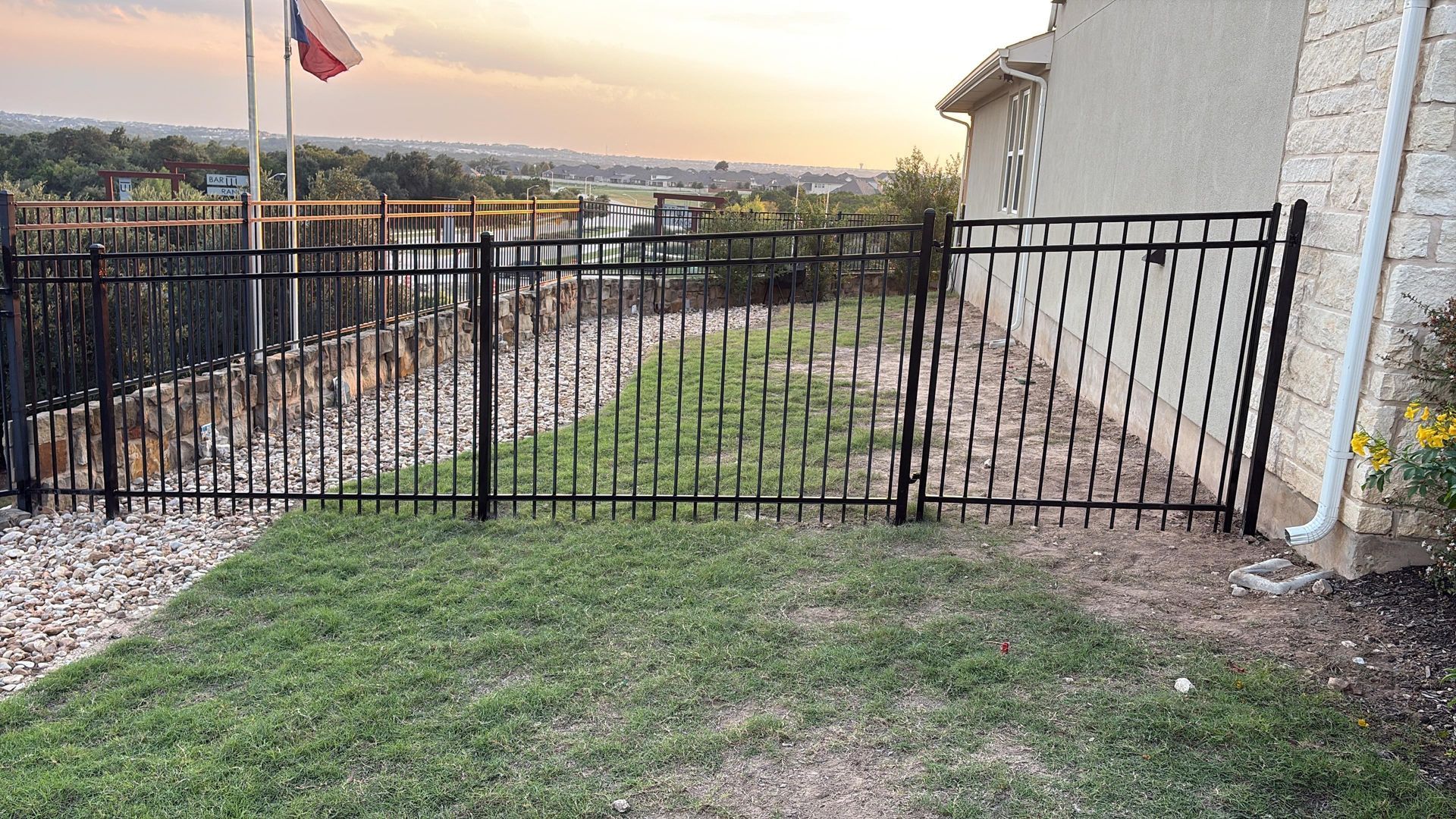 Black metal fence with a gate, green grass, and view of a cityscape at dusk.