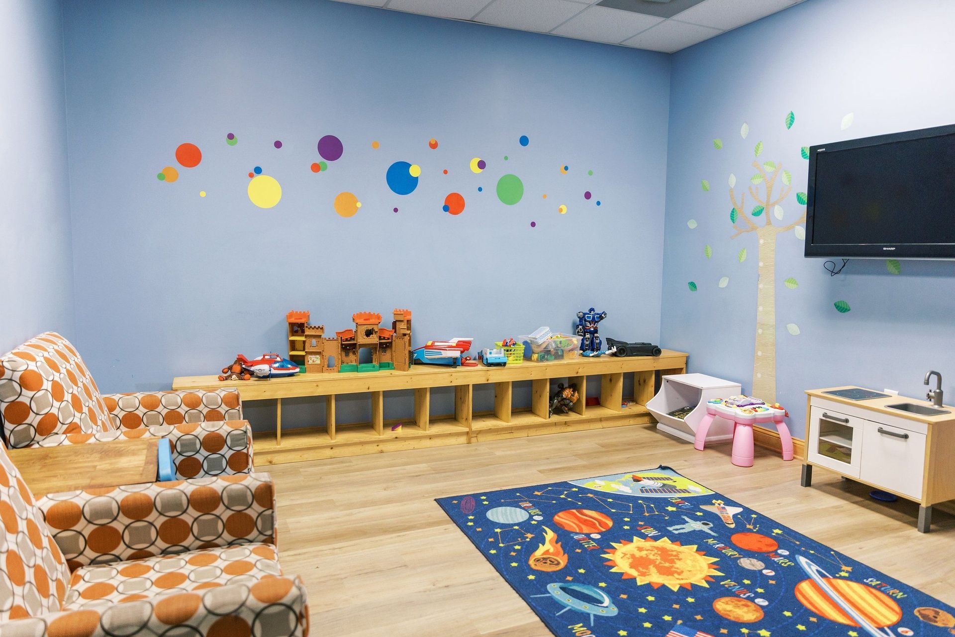 A play room with a rug , chairs , a table and a television.