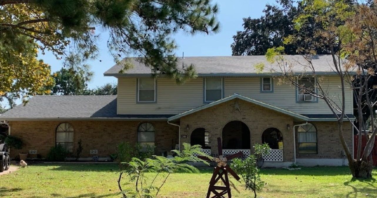 A large brick house with a gray roof is surrounded by trees and grass.