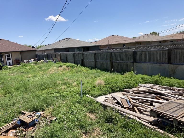 A fenced in yard with a pile of wood in the foreground and houses in the background.