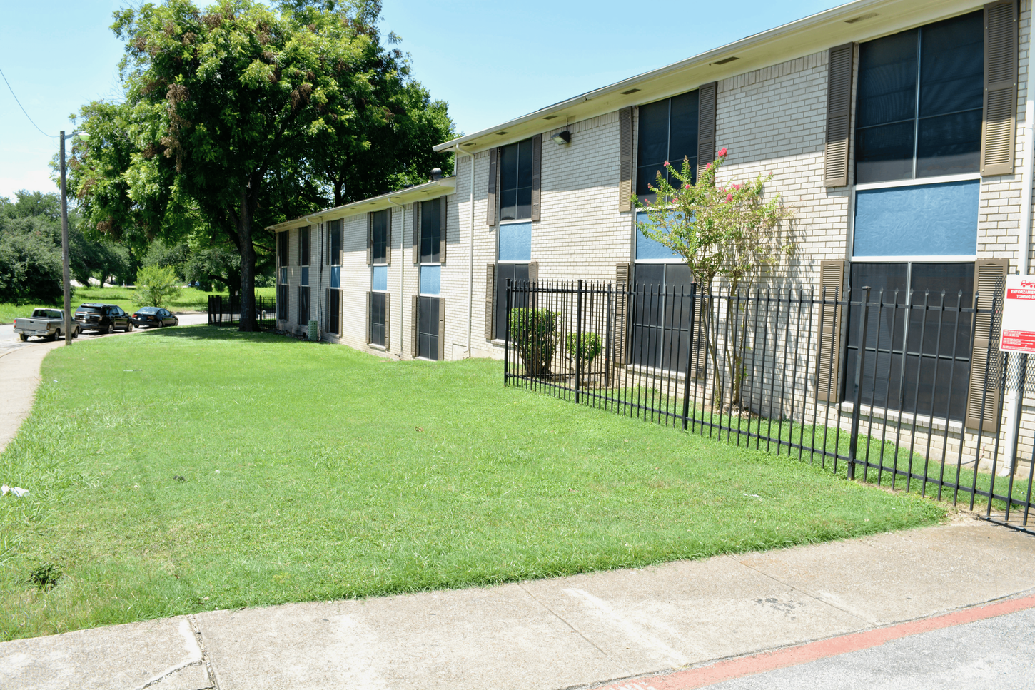 Two-story apartment building with light brick and dark-tinted windows; green lawn and sidewalk in front.