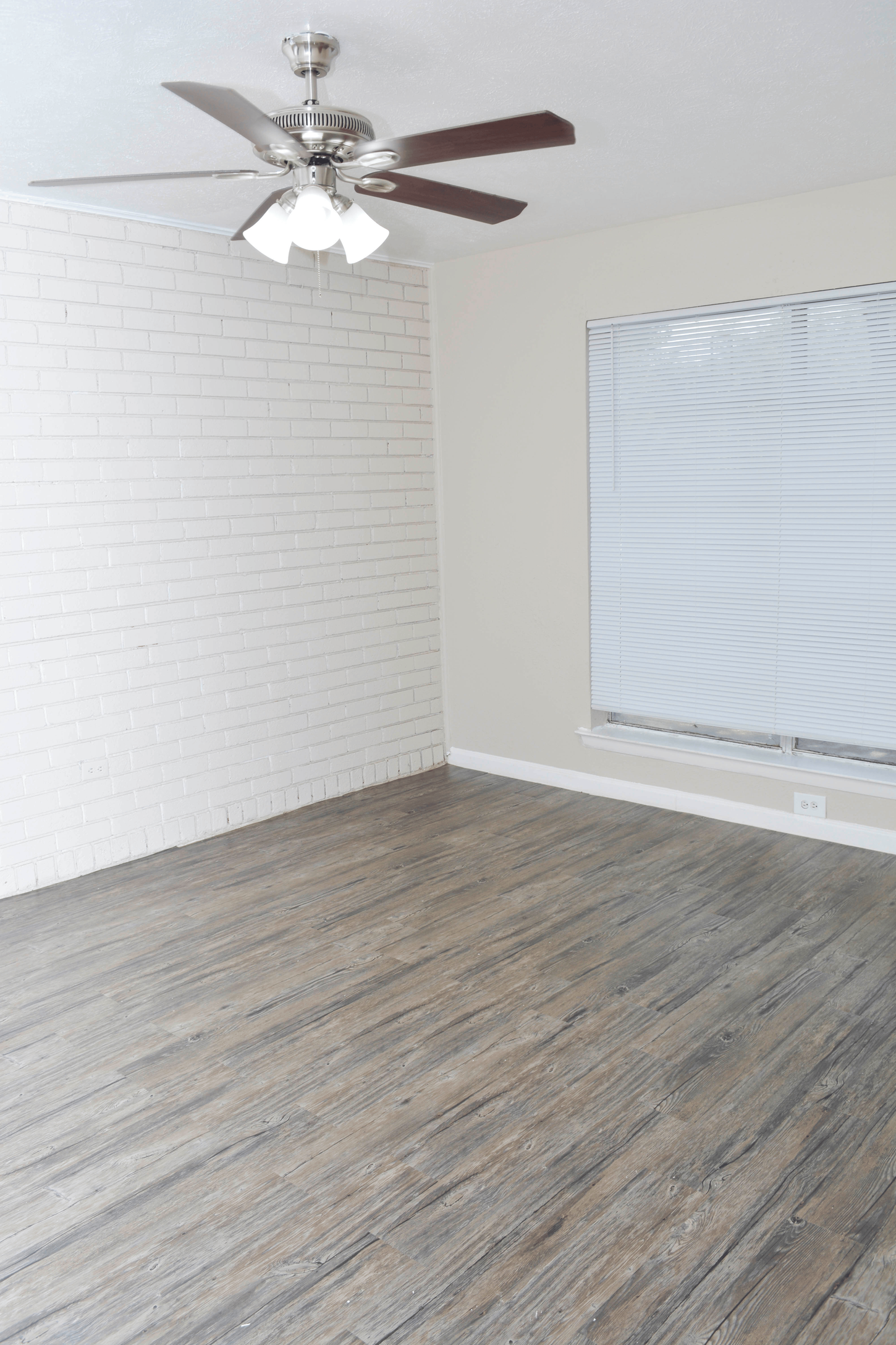 Empty room with hardwood-style floor, white brick wall, window with blinds, and ceiling fan with lights.