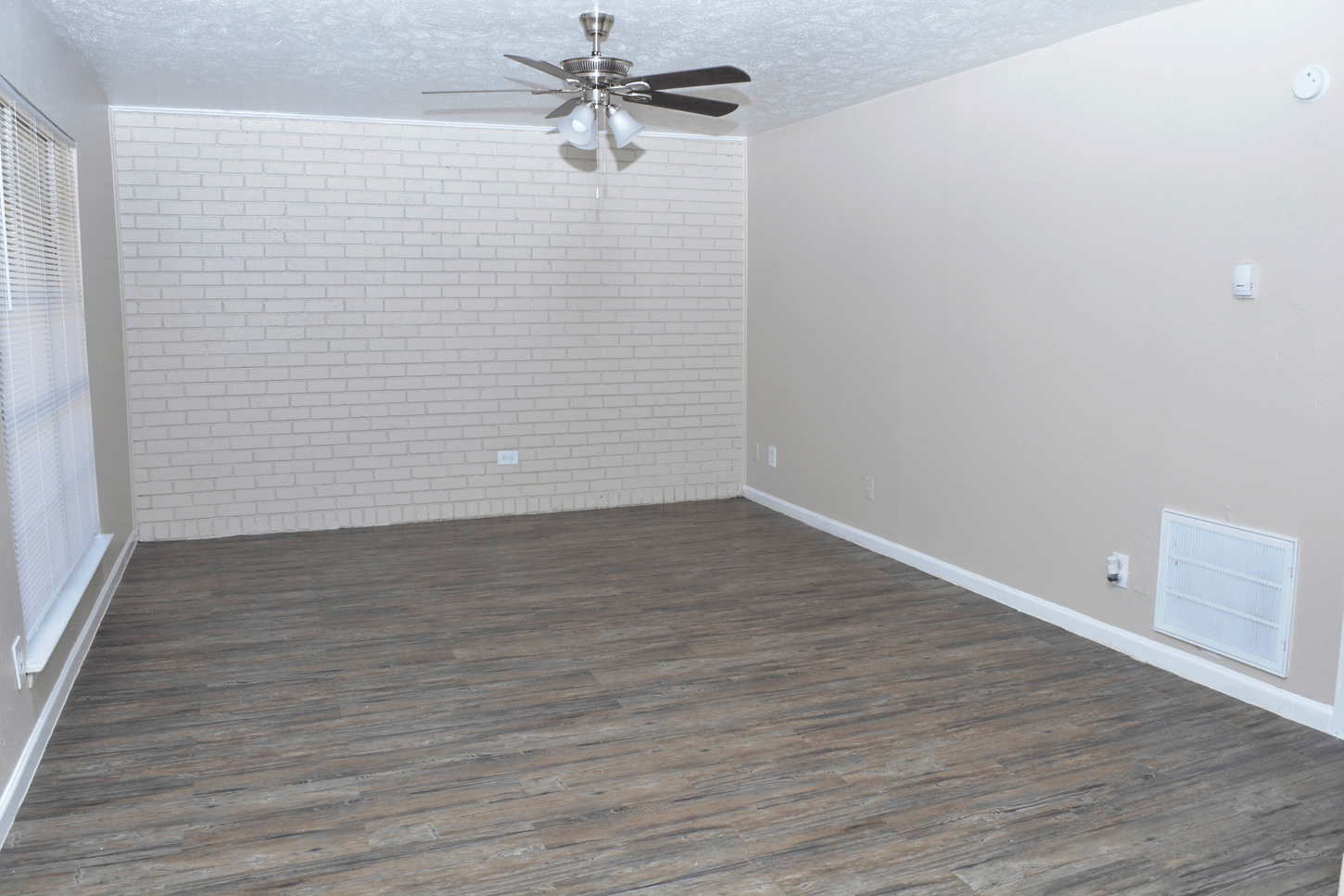 Empty living room with gray flooring, beige walls, a brick accent wall, and a ceiling fan.