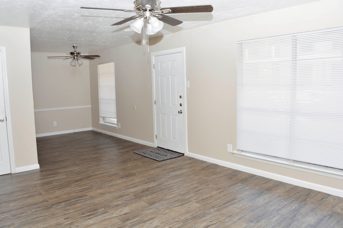 A living room with light brown flooring, white door, and two ceiling fans; beige walls.