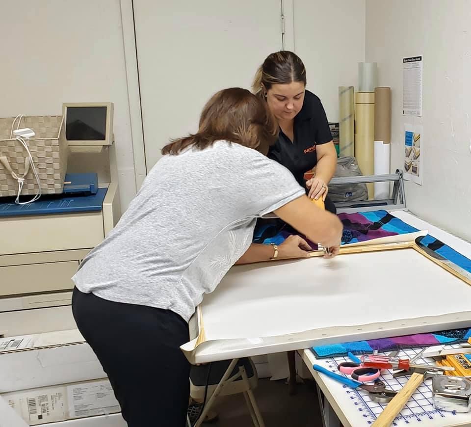 Two women stretching canvas over a wooden frame in a workshop.