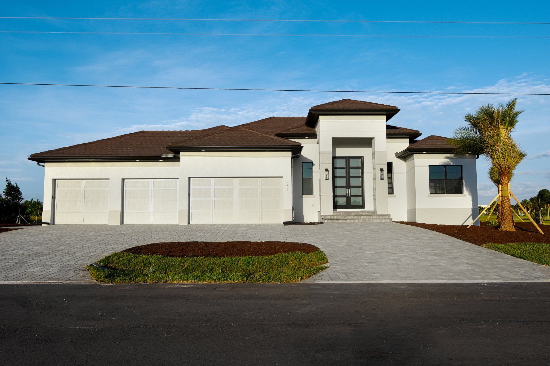 Modern, white house with three-car garage and blue sky. Gray paver driveway and palm tree.