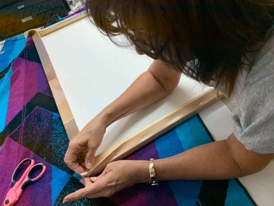 Woman stretching fabric over a wooden frame, using her hands. Scissors and colorful fabric are nearby.