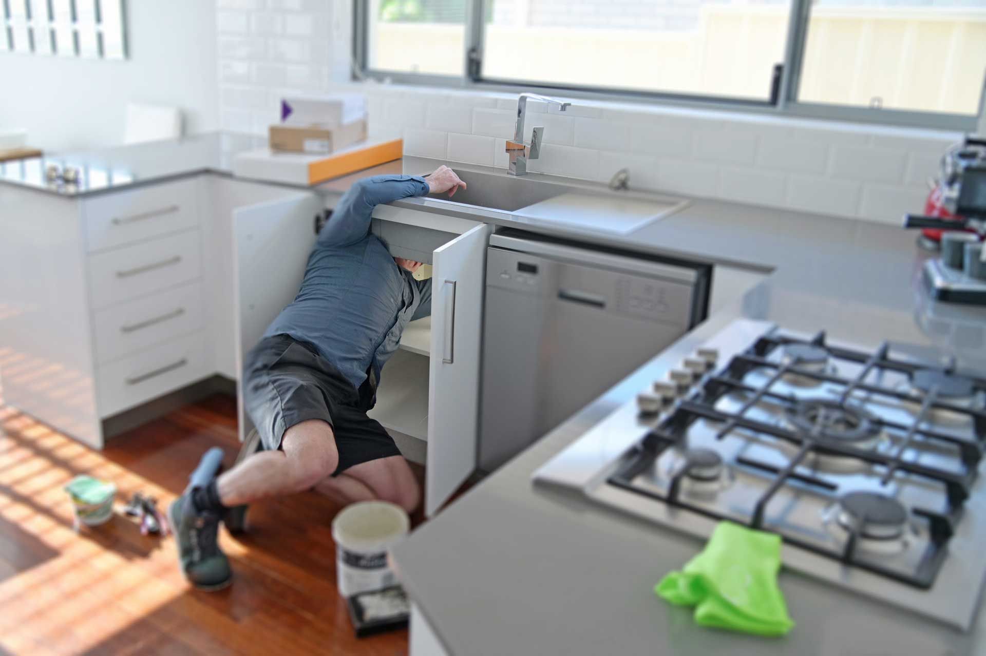 Plumber repairing kitchen sink cabinet in modern home interior setting.