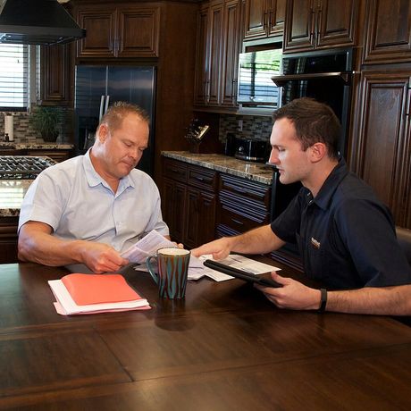Two men are sitting at a table in a kitchen looking at papers