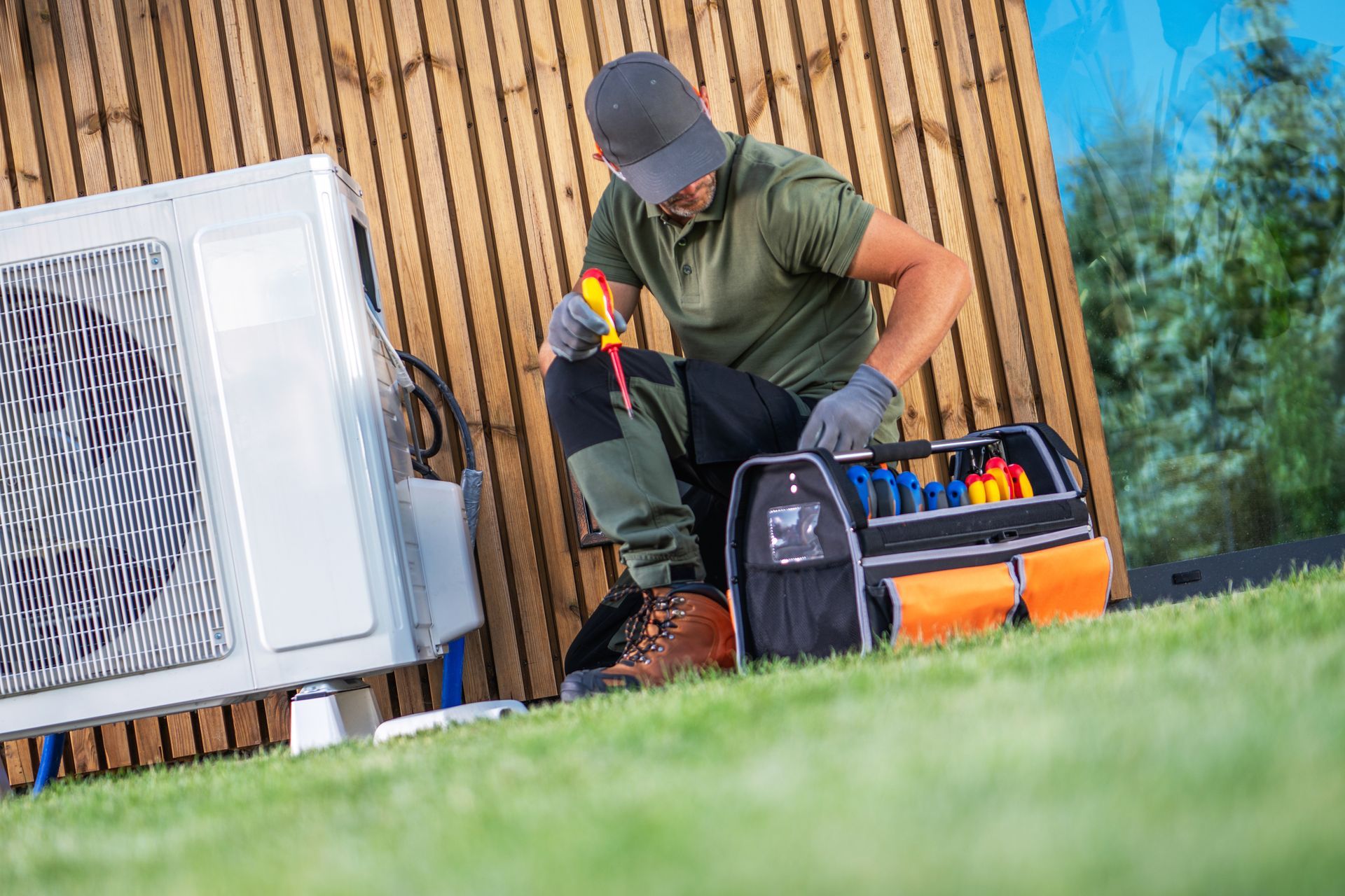 A technician in a cap and work clothes kneels on grass, using a tool to repair an outdoor air conditioning unit.