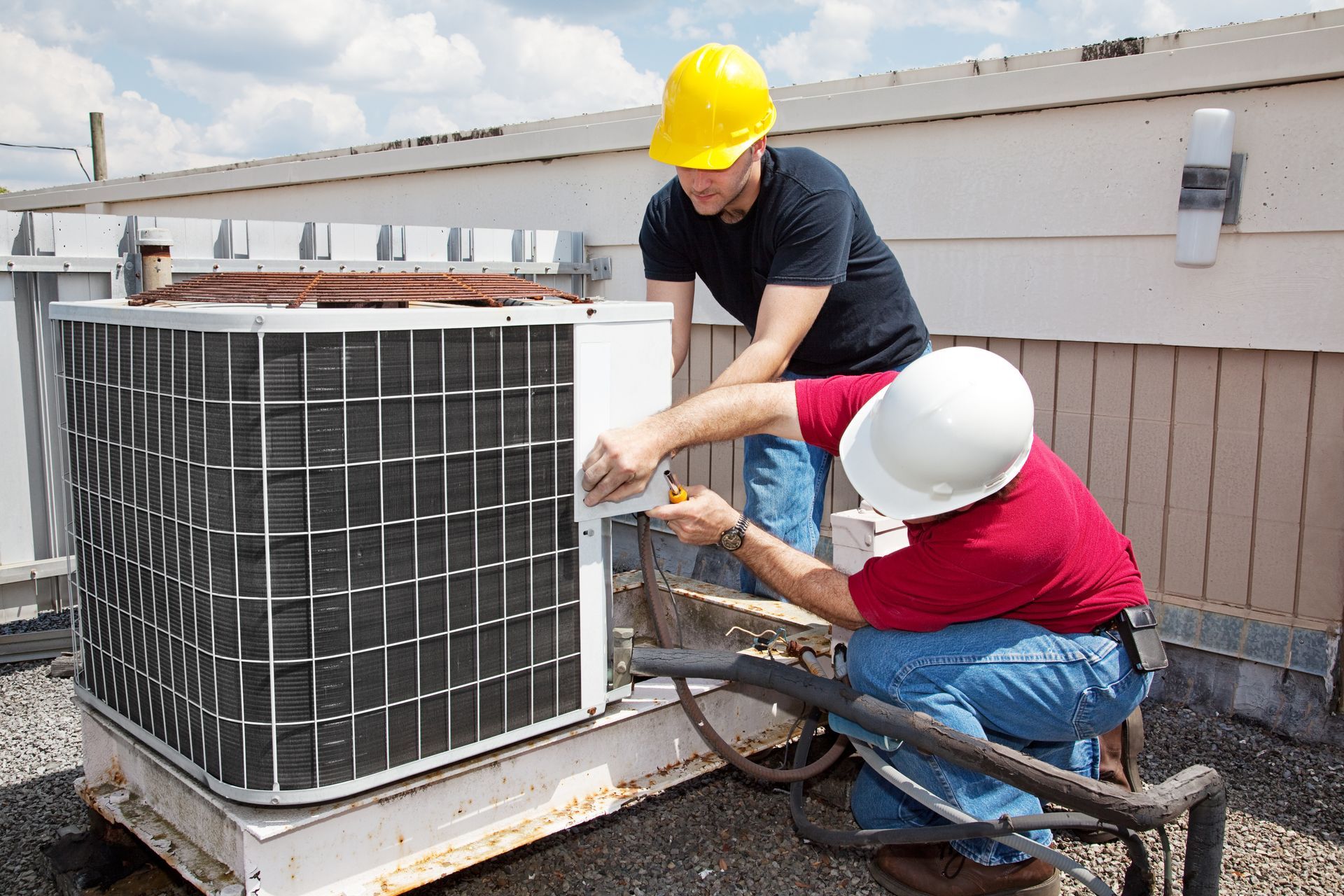 Two technicians in hard hats repair an outdoor HVAC unit on a rooftop.