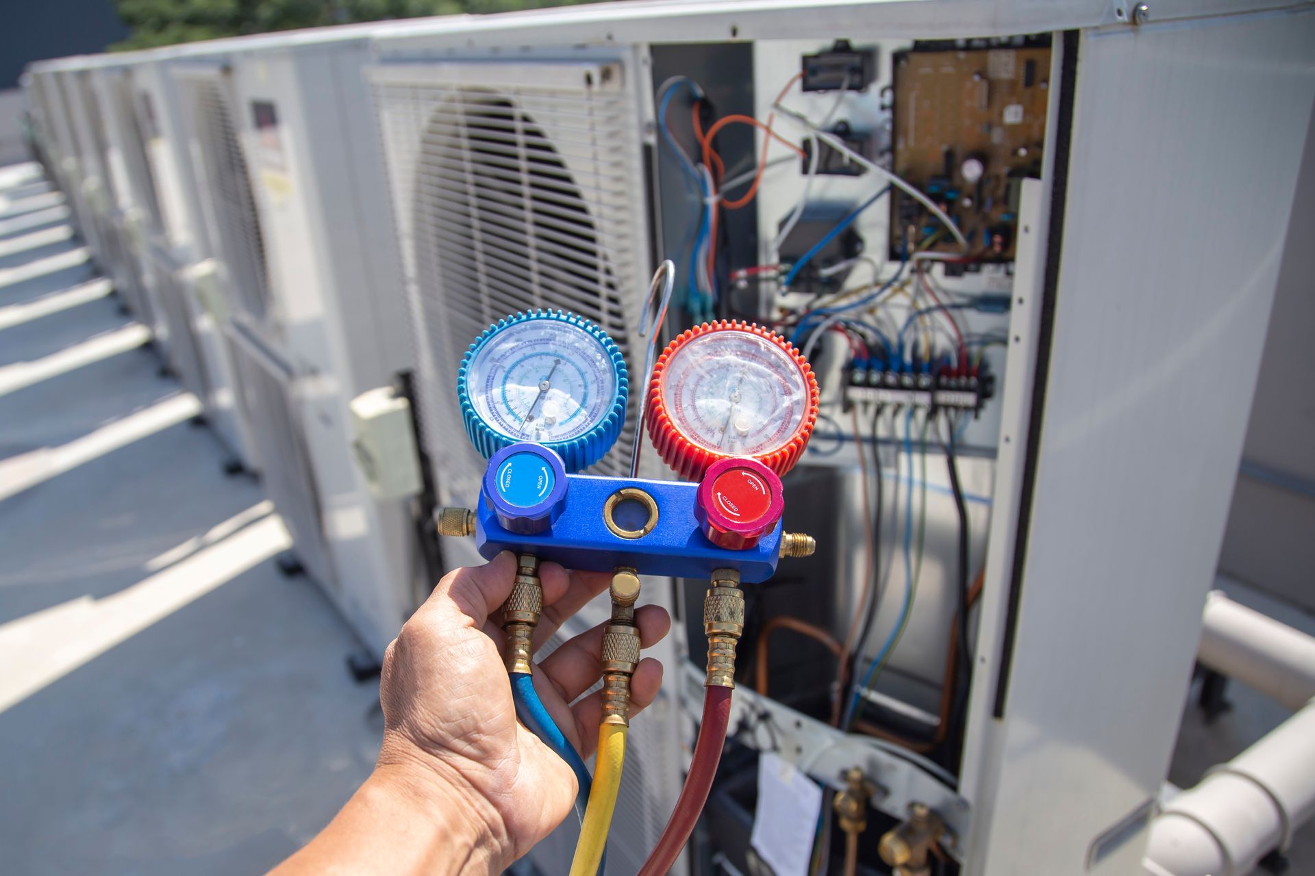 A hand holds an HVAC manifold gauge set while servicing an outdoor air conditioning unit.