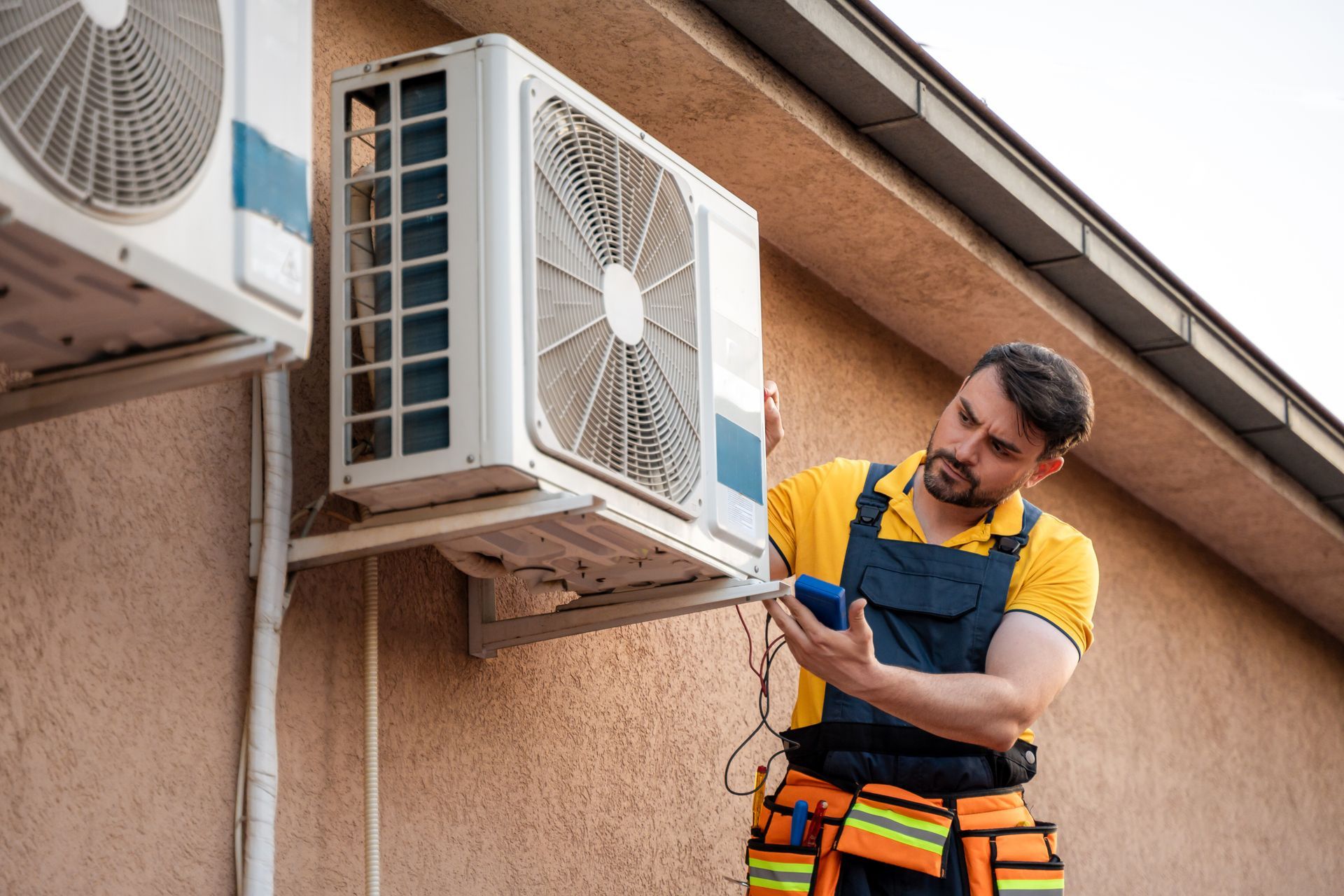 A technician in a yellow shirt and work overalls checks an outdoor air conditioning unit mounted on a tan wall.