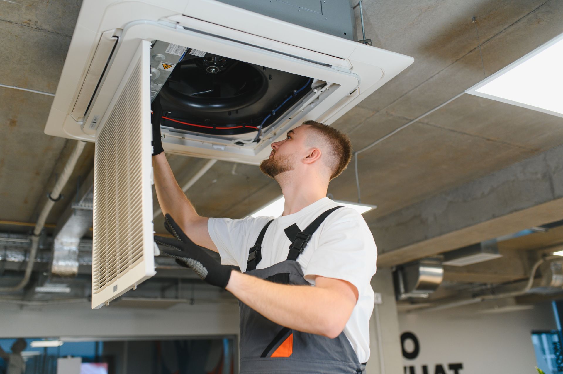 A technician in work coveralls and gloves services a ceiling-mounted air conditioning unit in an office setting.