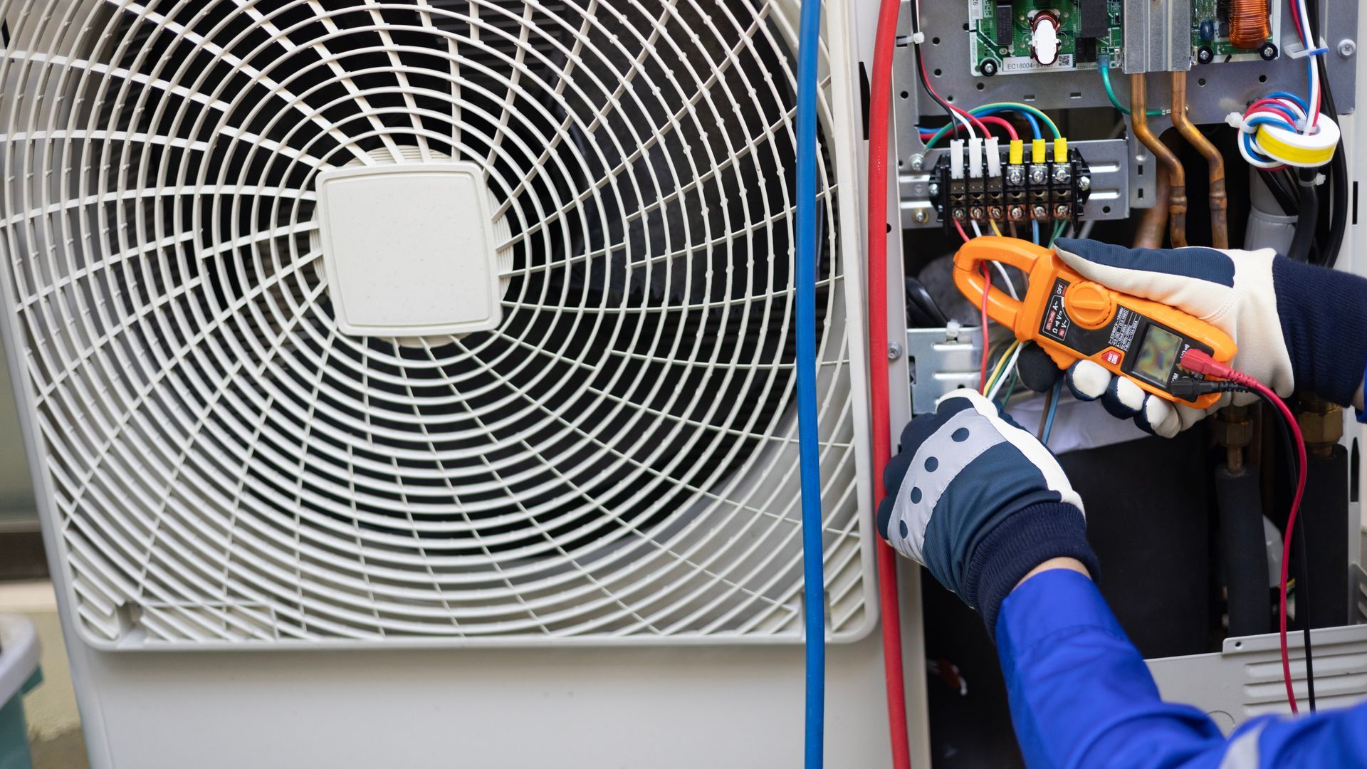 A person wearing work gloves uses a clamp meter to test electrical wiring inside an outdoor air conditioning unit.