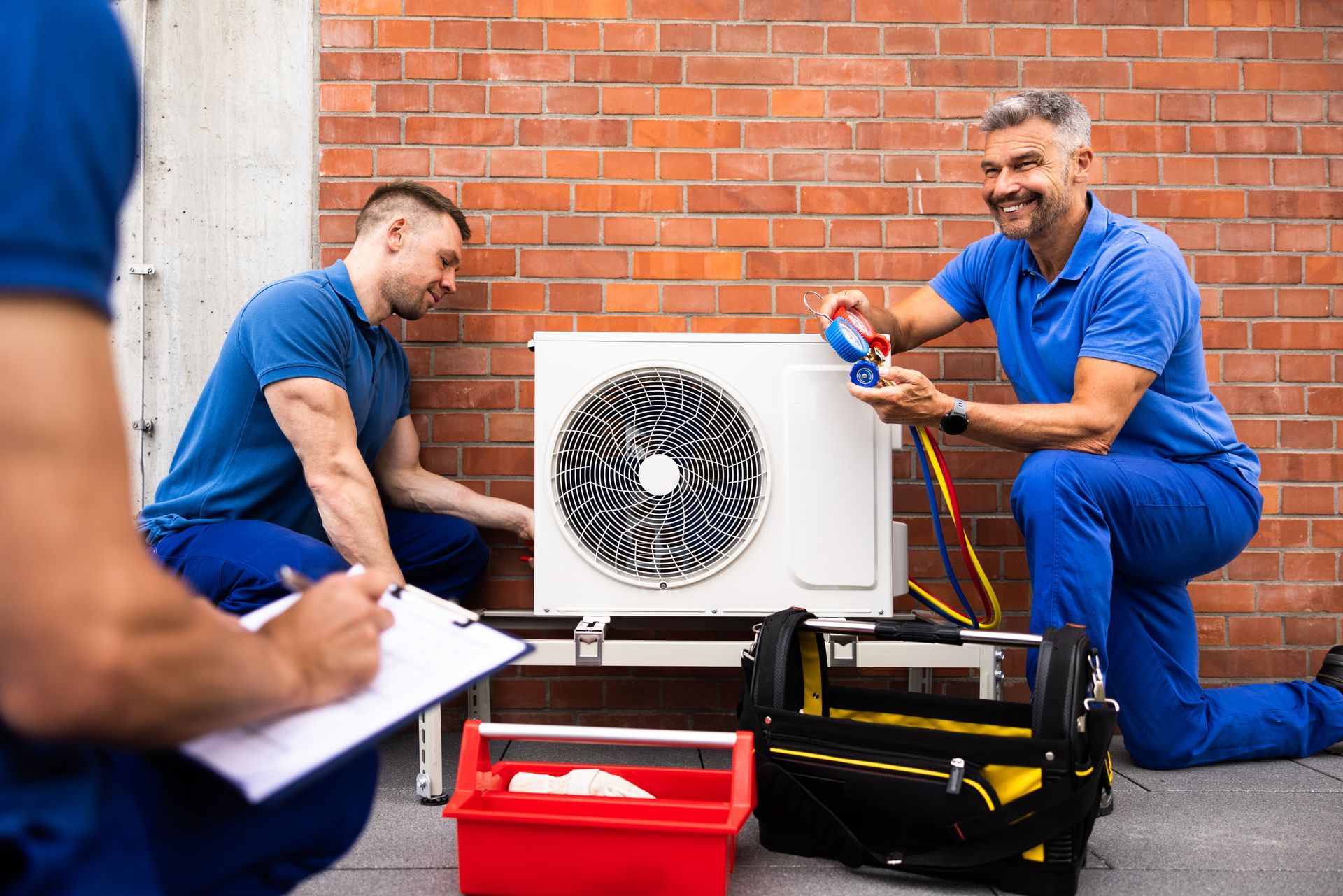 Technicians in blue uniforms inspect an outdoor HVAC unit against a brick wall, using tools and checking a clipboard.