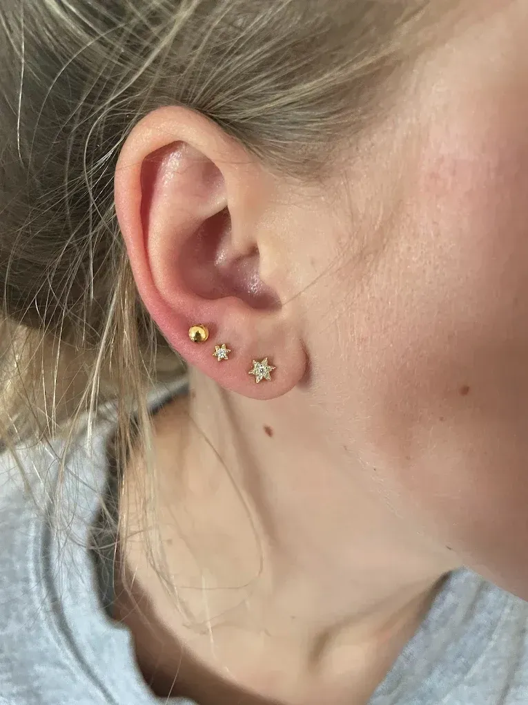 Close-up of a person's ear with three gold earrings: a stud, a star, and a small flower.