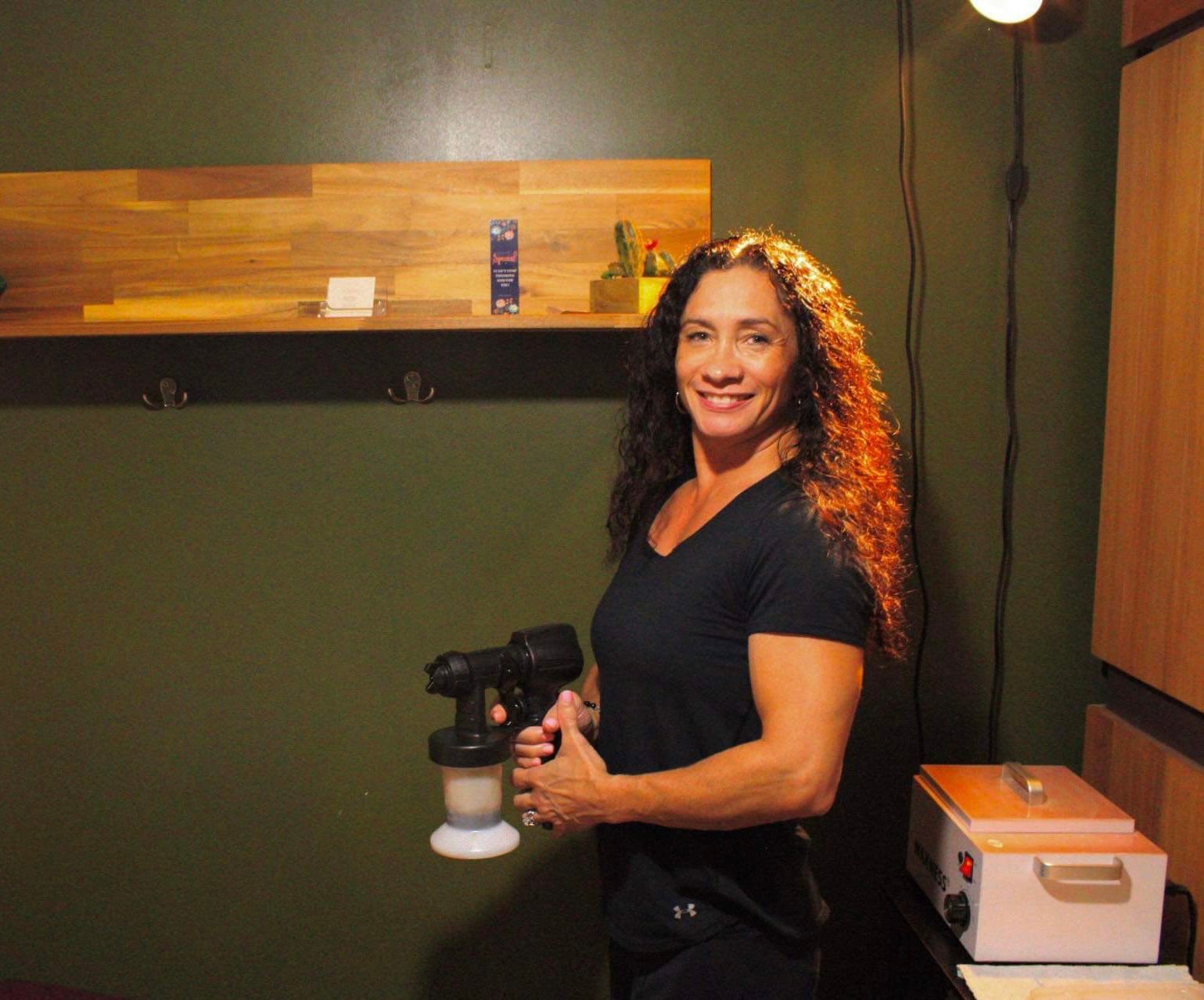 Woman holding spray tan gun, smiling. Green wall, wooden shelf, small heater in background.