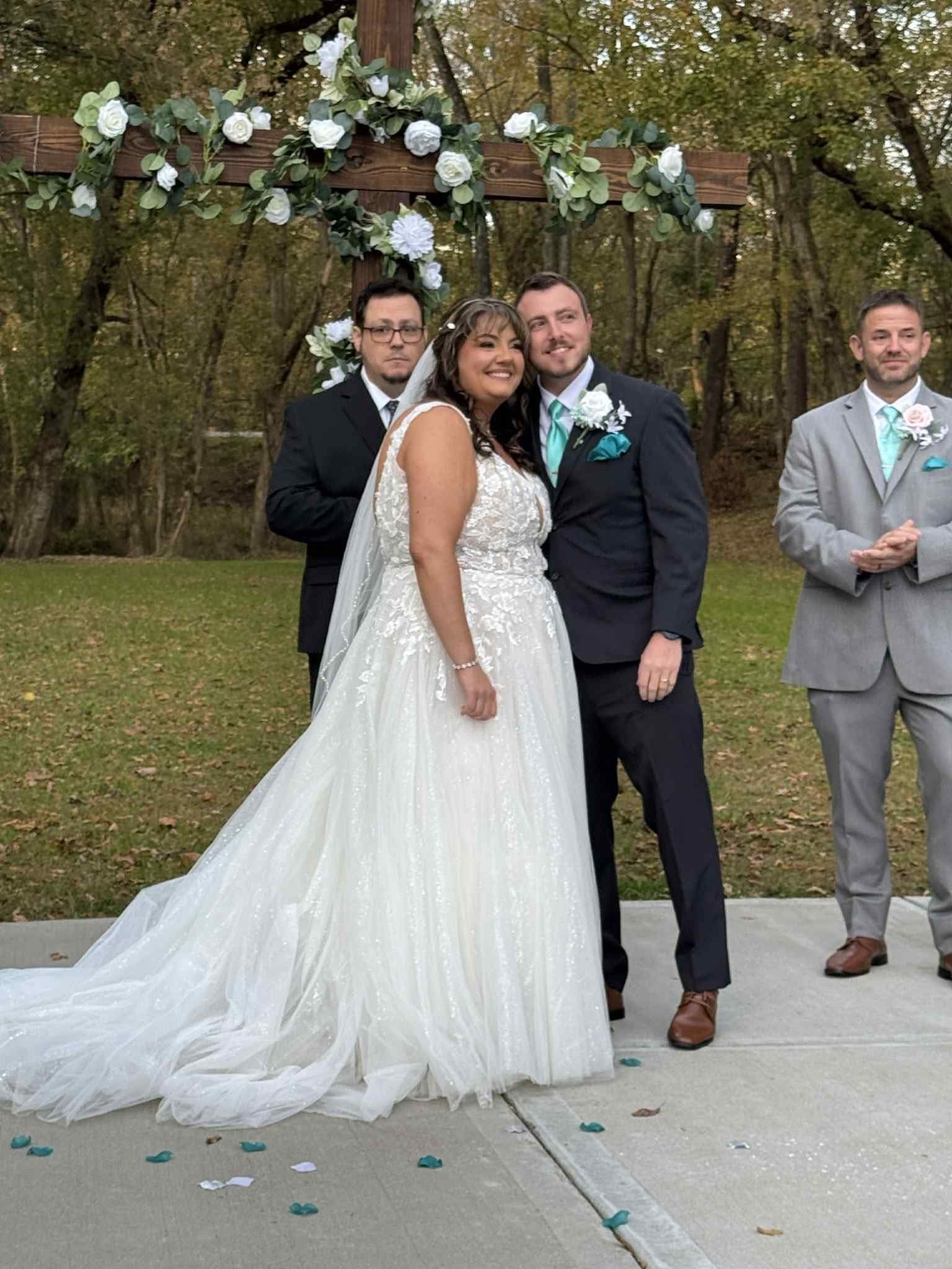 Couple stands, smiling, under a floral cross during their wedding ceremony. Two men in suits look on.